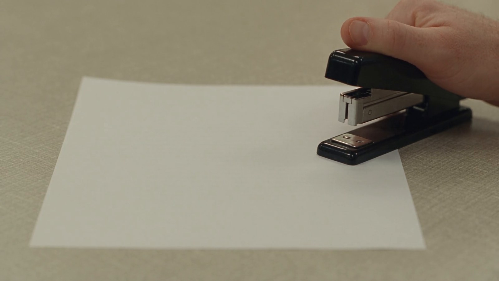 A hand presses an empty stapler onto a blank sheet of paper on a beige desk.
