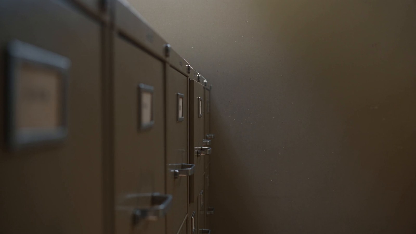 A row of beige filing cabinets in a dim office; one drawer has slid open an inch.